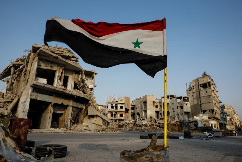 Una edificación destruida en el fondo y en el centro de la imagen la bandera de Siria. Los patrocinadores del terrorismo según Estados Unidos